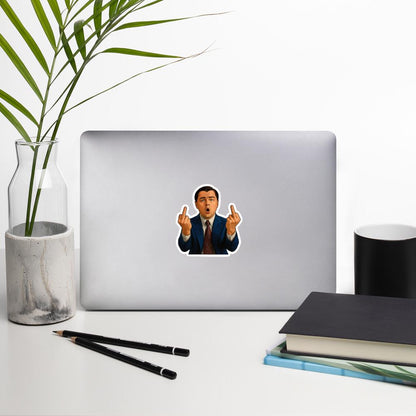 Laptop with a sticker of Jordan Belfort from the Wolf of Wall Street movie, in a suit with his middle fingers upwards, a white desk with a plant, pencils, and books.