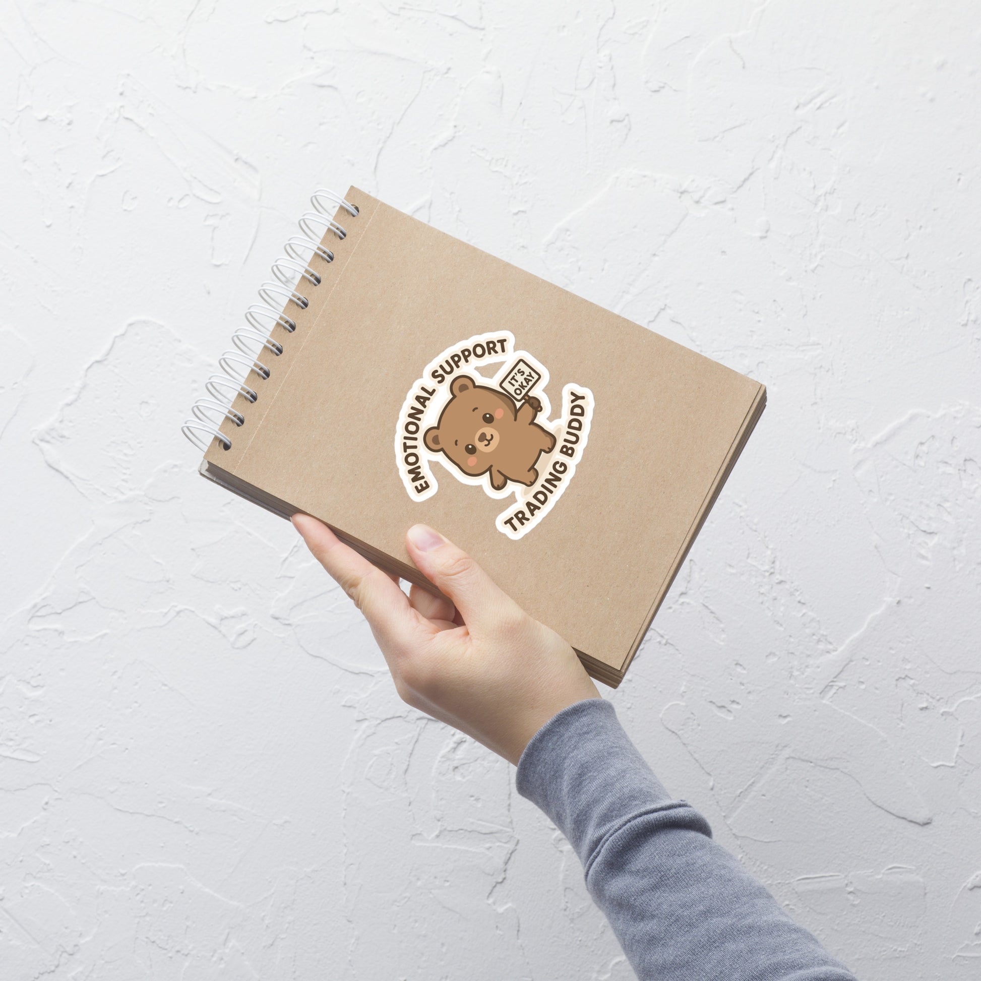 Brown notebook with a bear sticker labeled 'Emotional Support Trading Buddy' held by a hand on a light gray background